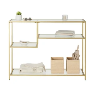 Gold metal-and-glass shelving unit with decor: plant, sunglasses, candle, and stacked books on top shelf; woven baskets and folded textiles on lower shelves.